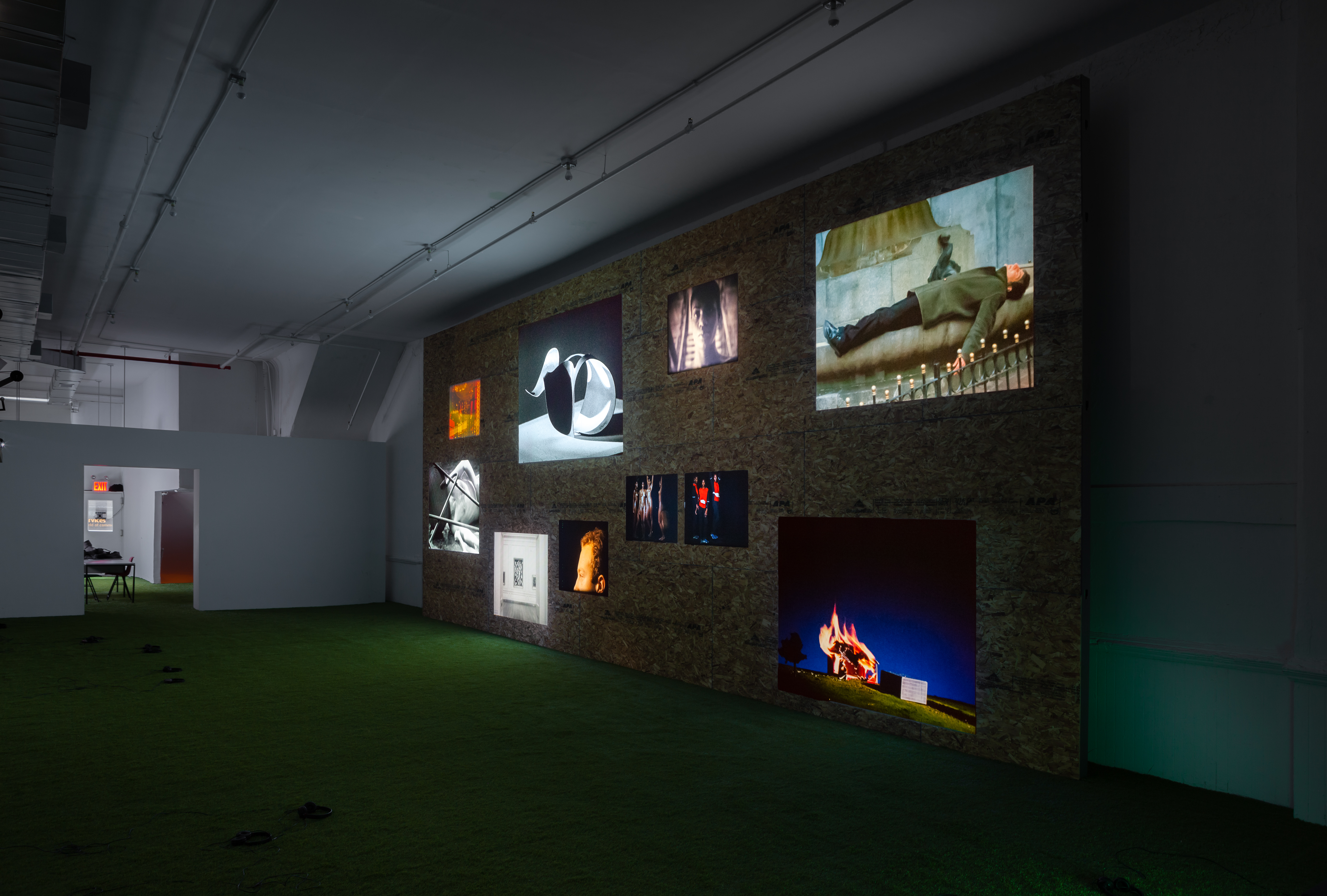 An angled view of scattered video projection stills at various heights and sizes on a wooden wall. The wooden wall is displayed in a room with white walls and faux-grass flooring.