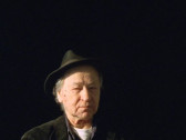 A portrait of Jonas Mekas in Rembrandt lighting looking directly into the camera, wearing his signature fedora and standing against a black background.