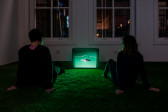 Two people seated with their legs outstretched on a green carpet in front of a small television monitor, wearing headphones.