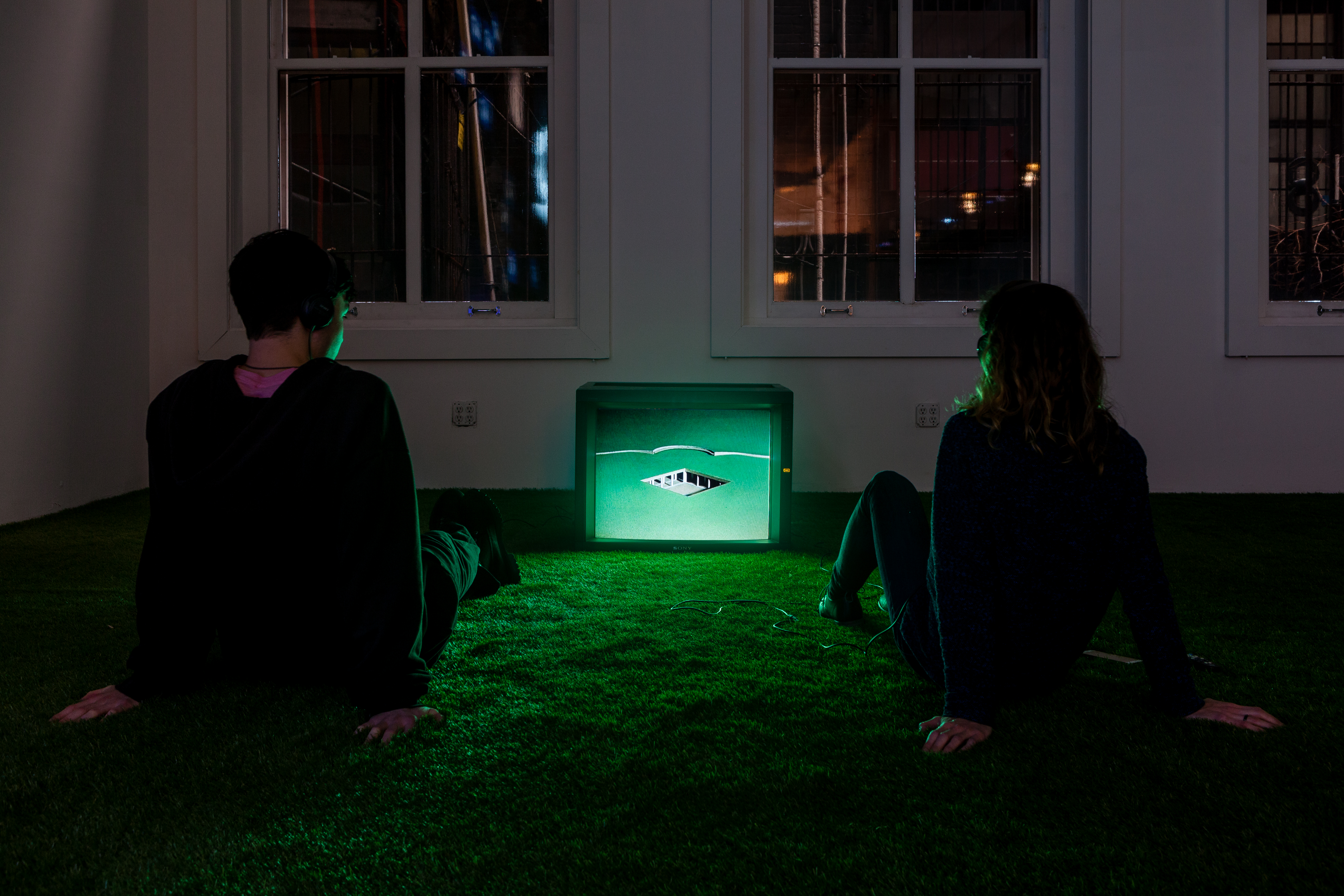 Two people seated with their legs outstretched on a green carpet in front of a small television monitor, wearing headphones.