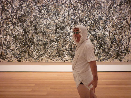 A man is shown in front of a Jackson Pollock painting wearing a frilly baby bonnet, sunglasses, a pacifier, a white t-shirt and a white cloth that resembles a diaper.