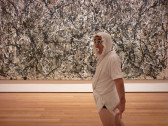 A man is shown in front of a Jackson Pollock painting wearing a frilly baby bonnet, sunglasses, a pacifier, a white t-shirt and a white cloth that resembles a diaper.