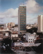 Photographic print of an urban skyline on a sunny day. A small skyscraper juts out from a cluster of lower buildings at the image's center. Text emblazoned across the facade of a low, white building in front of the skyscraper reads, 