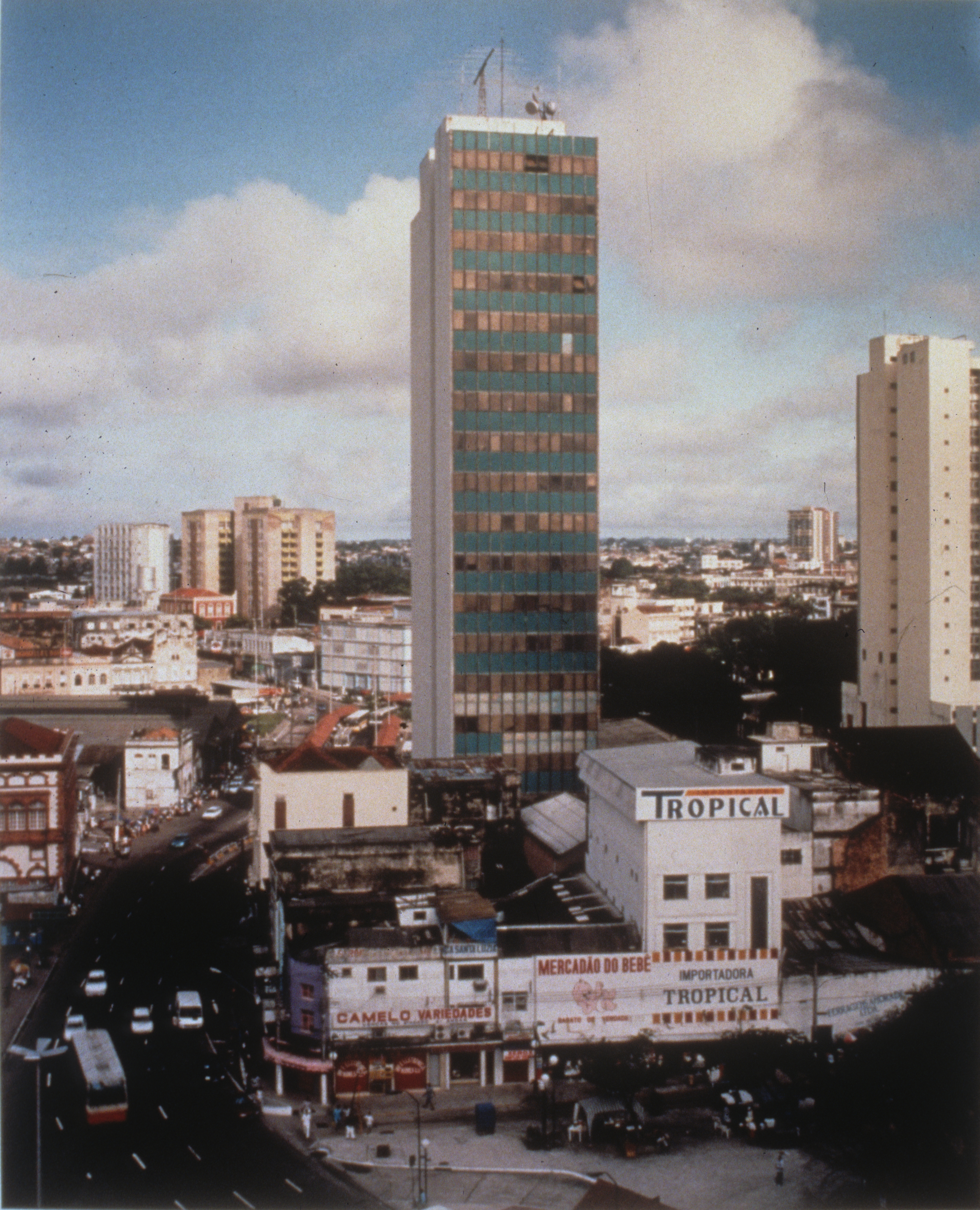 Photographic print of an urban skyline on a sunny day. A small skyscraper juts out from a cluster of lower buildings at the image's center. Text emblazoned across the facade of a low, white building in front of the skyscraper reads, 