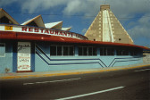 Photgraphic print of a low, blue and red building situated next to an empty multi-lane road on a sunny day. The building has a low blue facade with many windows, which face the street, and is decorated with two angular blue stripes. The building has a low, curved red roof. Text running along the side of the roof reads, 