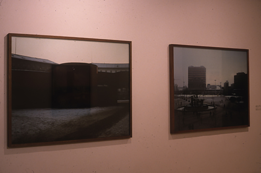 Two photographs displayed side-by-side on a white gallery wall. The photographs are of identical size and are installed in thin, dark frames. The photograph on the left depicts a nondescript industrial building, while a tall urban building is visible in the skyline of the image to the right.
