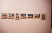 Seven small, color photographic prints installed in light wooden frames on a white gallery wall. The photographs are displayed side-by-side in a horizontal line, some displayed in a landscape and some in a portrait orientation. All display images of various buildings in sunny environments.