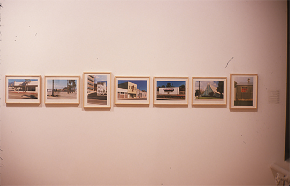 Seven small, color photographic prints installed in light wooden frames on a white gallery wall. The photographs are displayed side-by-side in a horizontal line, some displayed in a landscape and some in a portrait orientation. All display images of various buildings in sunny environments.