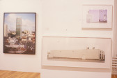 Three framed photographs installed in a gallery space with white walls and wooden flooring. Two lighter photographs of white, industrial buildings are installed one above the other on a freestanding wall on the image's left side. Visible behind and to the right of the freestanding wall is a third, color photograph of a city skyscraper rising out of an urban landscape.