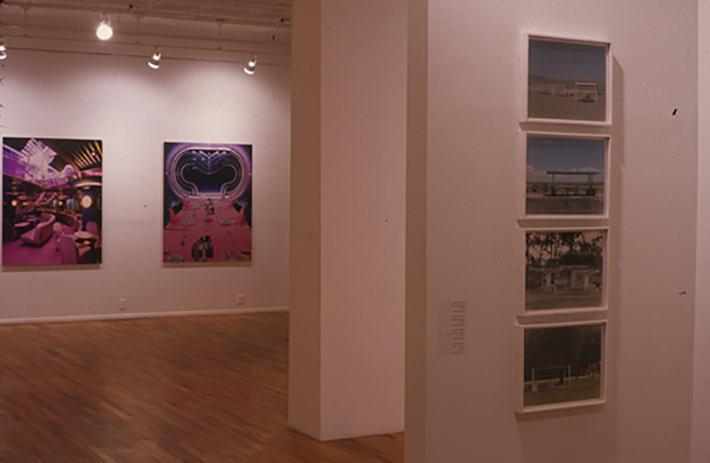Several photographs installed in a gallery space with white walls and wooden flooring. Four color photographs are arranged in a vertical line on a wall to the right side of the image. The wall terminates to the left of the displayed photographs, and a second wall extends into the gallery space directly behind it. Two large images comprised of pink and purple hues hang on a far gallery wall in the background of the image. The image on the left displays a futuristic looking, interior space.