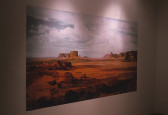 A dimly lit, color photograph of a desert installed on a white gallery wall. The desert ground is a bright orange color and includes many large rock features. The sky above is slightly blue and filled with clouds.