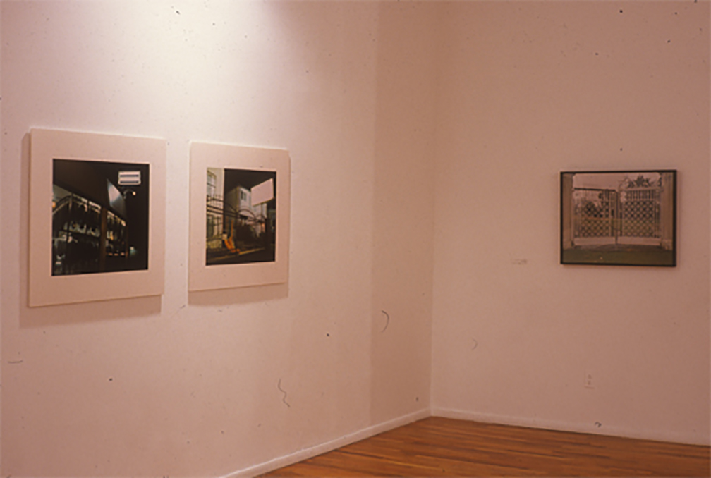 Three framed photographs installed in the corner of a gallery space with white walls and wooden flooring. Two darker, square photographs with large white frames are installed on the corner