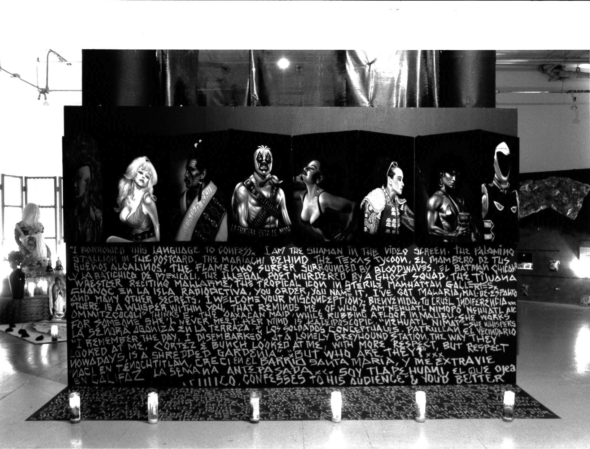 A folding wall is erected in the center of a gallery space.  Its top features painted portraits of eight people from the waist up.  Below them, the lower half of the wall is covered in white, handpainted text on a black background.   The text extends onto the floor, and its bottom edge is bordered by a row of votive candles.  Behind this wall, other artworks are partially visible.