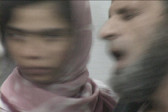 Two figures' faces positioned close to one another. The woman on the left looks away from the camera, while the man on the right closes his eyes and opens his mouth. The image is blurred, as though the figures are in motion.