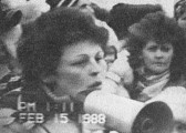 Black and white video still of a figure in a packed crowd, speaking into a microphone. A date and time stamp at the bottom left corner of the still read, 