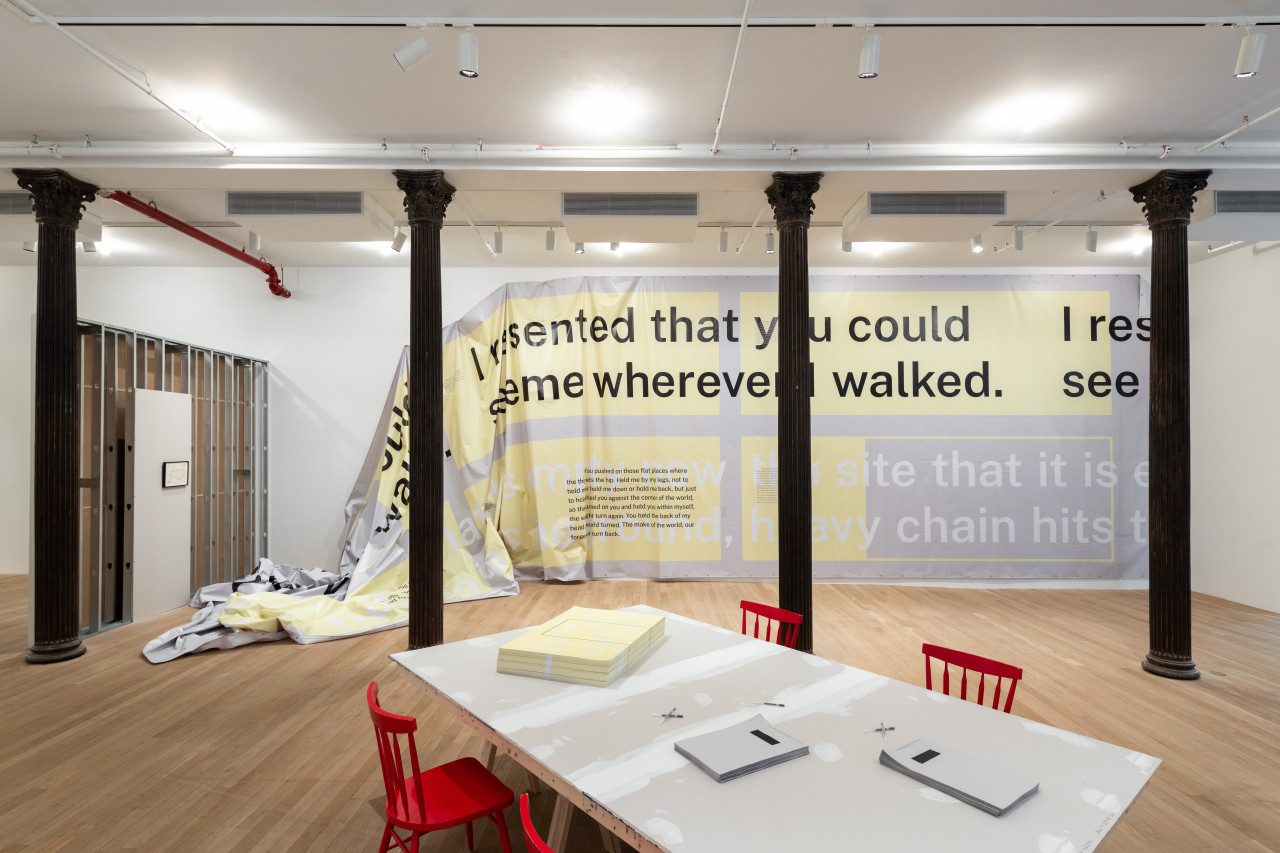 A color photograph of a gallery space. Behind three dark columns a large vinyl printed with text is attached to the wall, falling to the ground on the left side. In the center a large grey table with four red chairs and stacks of yellow and grey posters is visible.