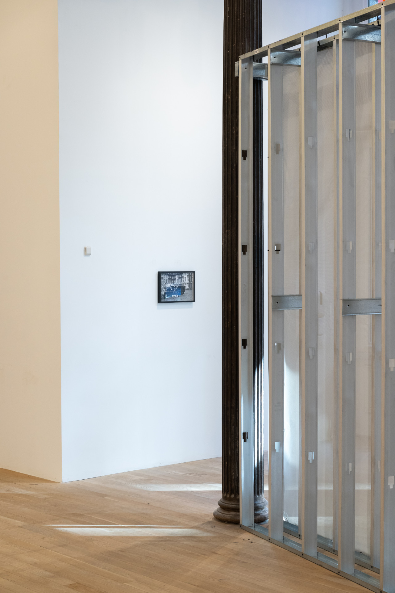 A color photograph of a gallery space. A small framed photograph hangs from the far wall, perpendicular to a dark metal column next to grey metal scaffolding.