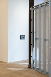 A color photograph of a gallery space. A small framed photograph hangs from the far wall, perpendicular to a dark metal column next to grey metal scaffolding.