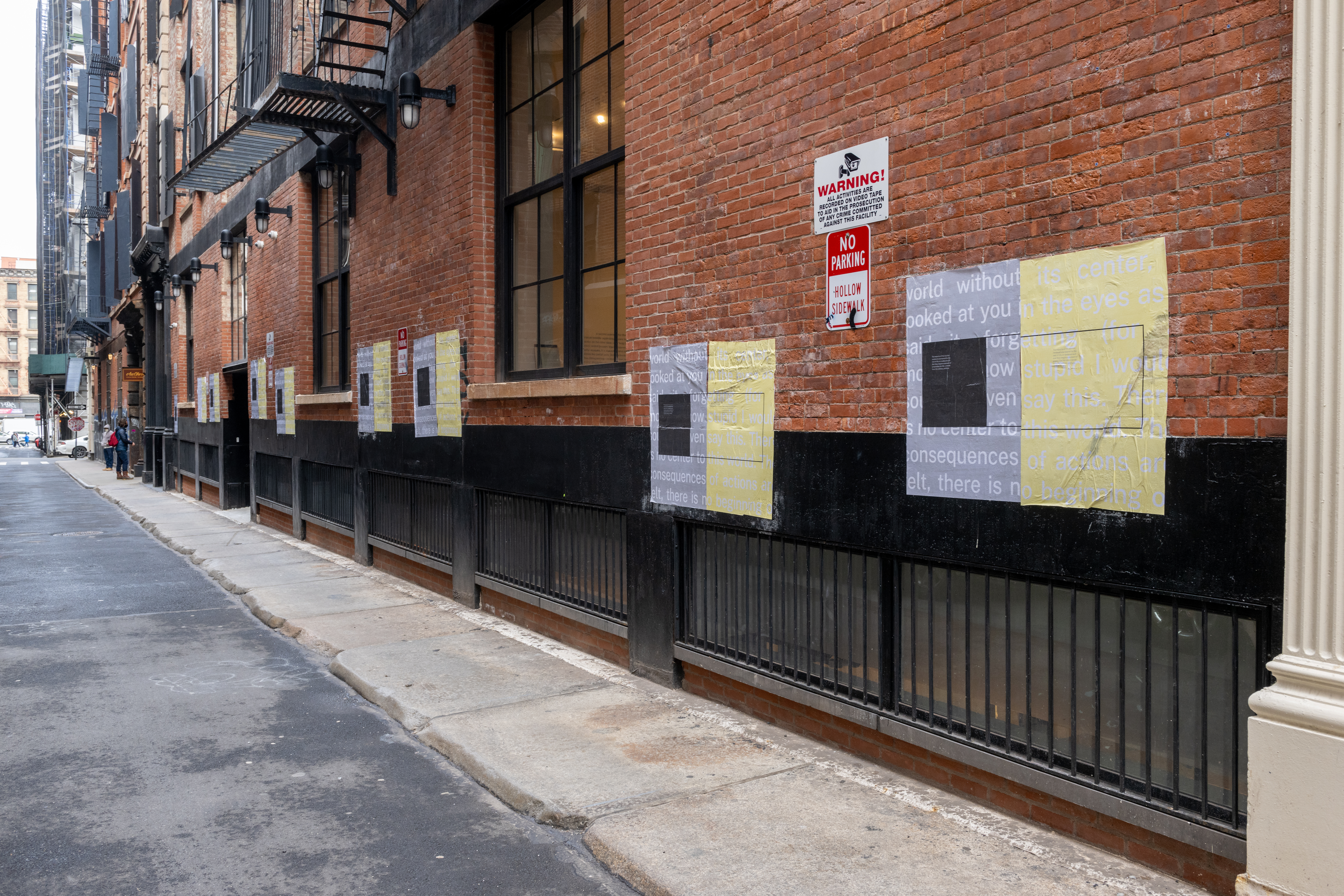A color photograph looking down an alleyway. Sets of grey and yellow posters hang from the brick wall of the alleyway.