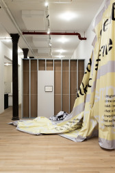A color installation photograph of a gallery space with a row of dark metal columns on the left, an exposed interior of a drywall with one grey panel in the center, and a large swath of partially hung grey and yellow vinyl on the right.