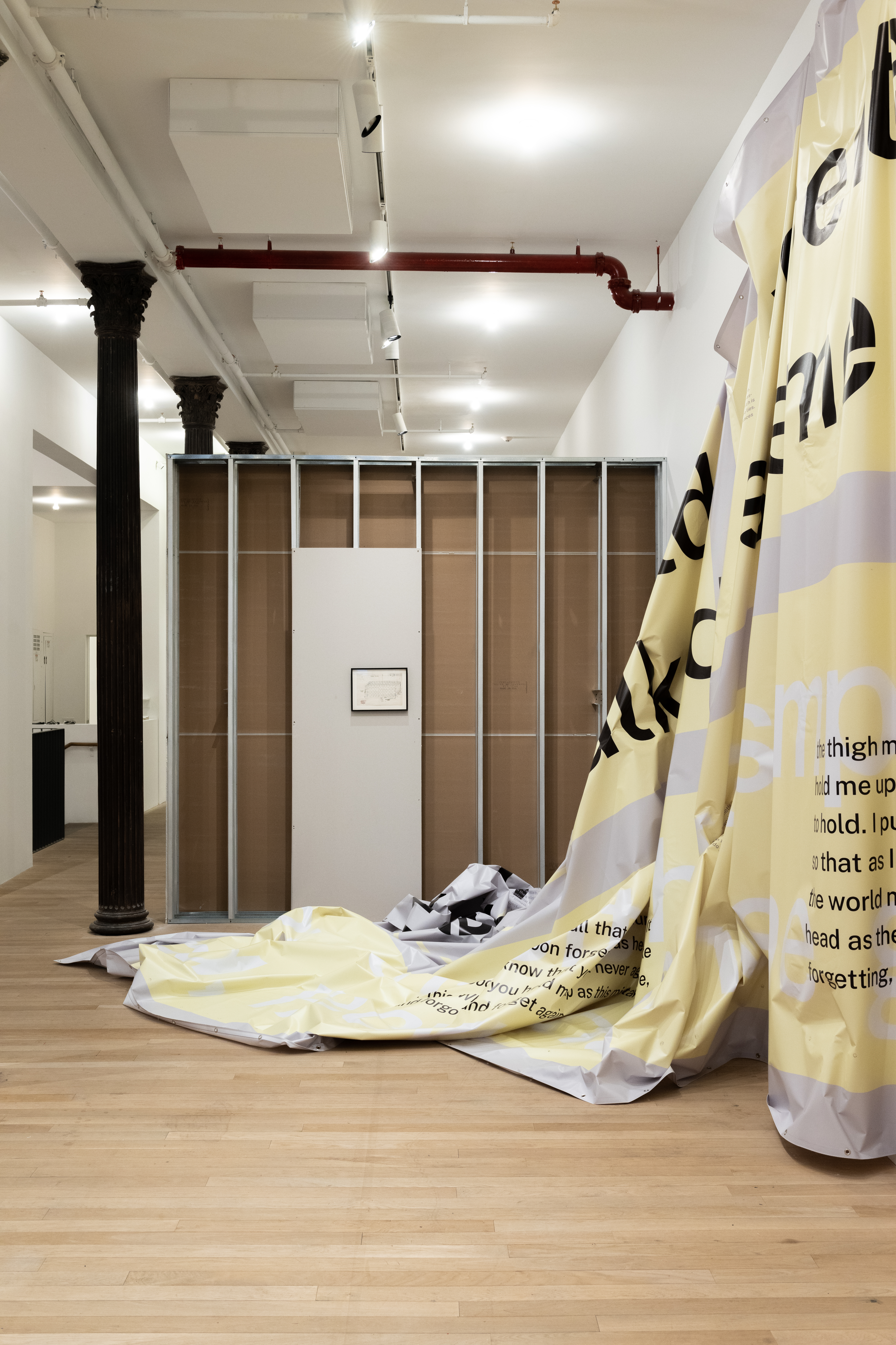 A color installation photograph of a gallery space with a row of dark metal columns on the left, an exposed interior of a drywall with one grey panel in the center, and a large swath of partially hung grey and yellow vinyl on the right.