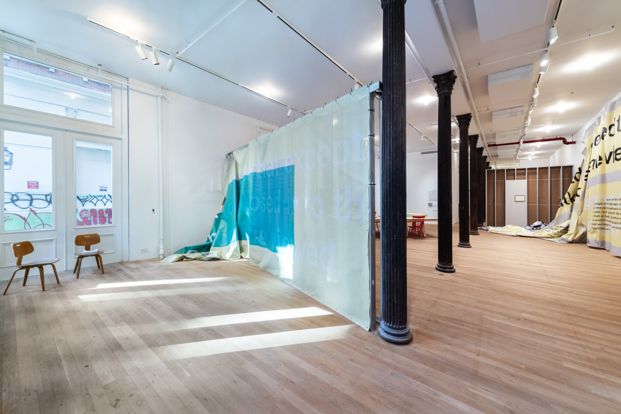 A color installation photograph of a gallery space bisected by a row of dark metal columns. Two wooden chairs are placed near a piece of semi-transparent grey and blue fabric partially hung from a grey metal scaffold. A large piece of grey and yellow vinyl hangs from the wall on the right.