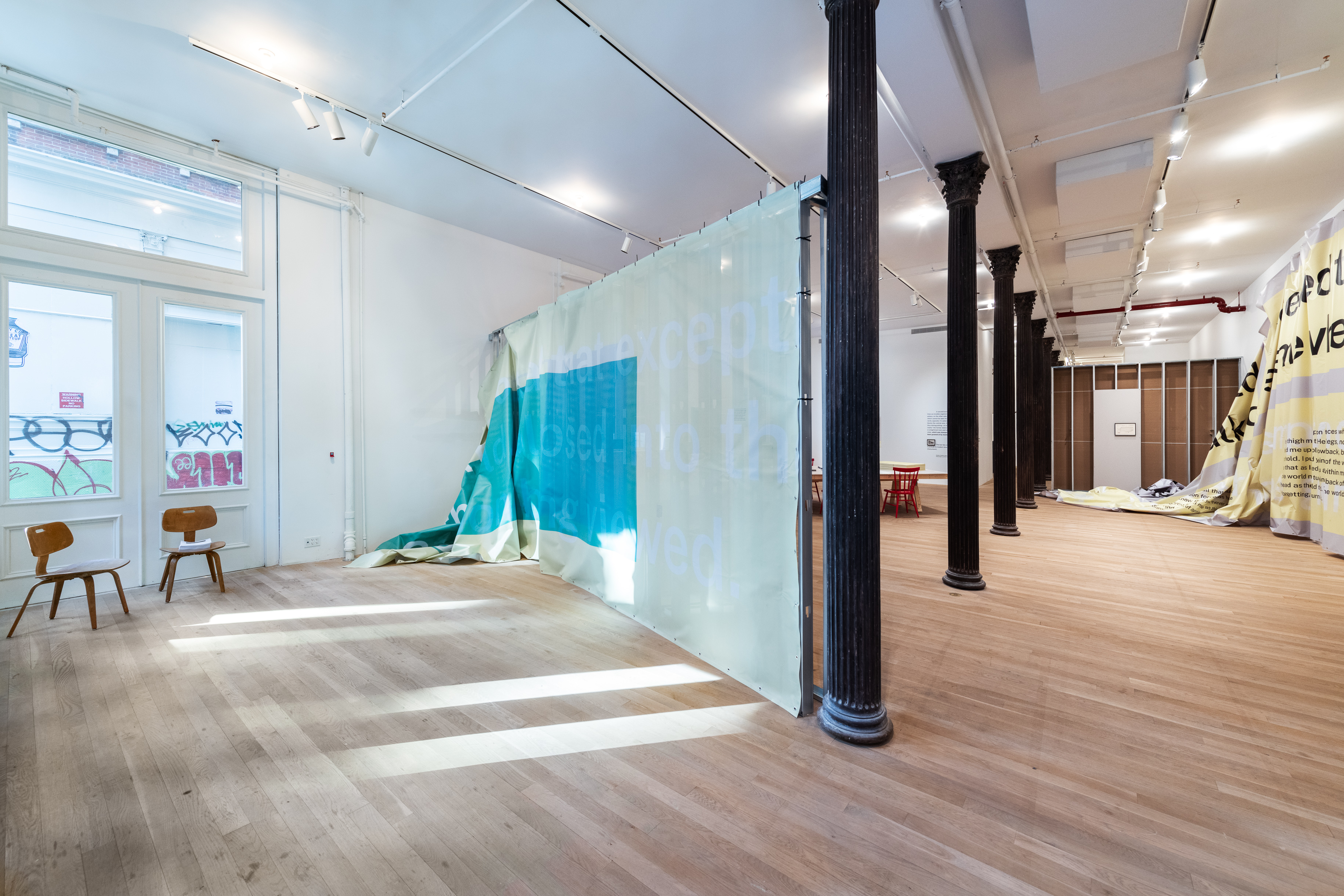 A color installation photograph of a gallery space bisected by a row of dark metal columns. Two wooden chairs are placed near a piece of semi-transparent grey and blue fabric partially hung from a grey metal scaffold. A large piece of grey and yellow vinyl hangs from the wall on the right.
