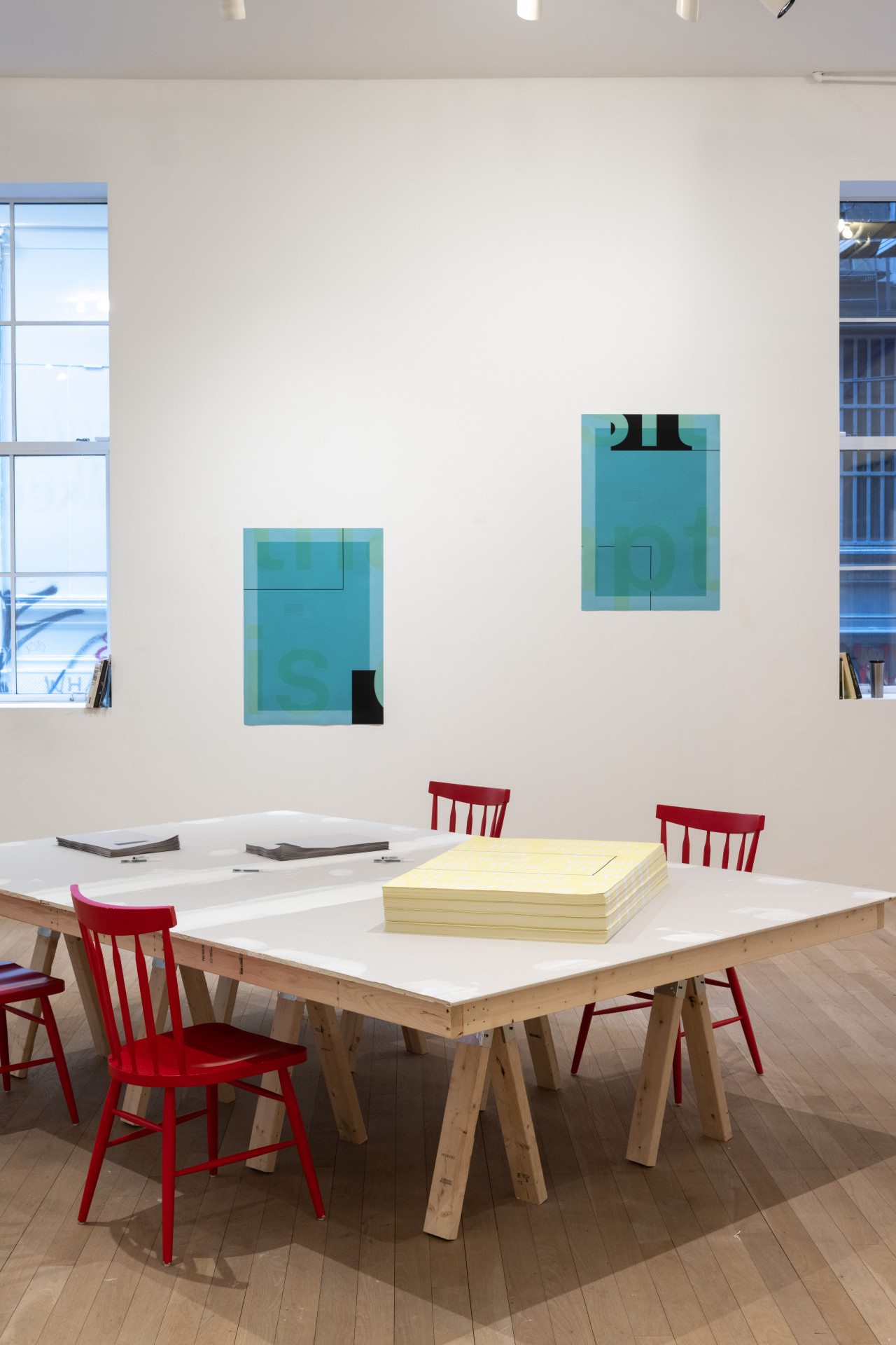 A color photograph of a gallery space with two partial views of windows. In between the windows, two blue posters are attached to the wall. Directly in front of the posters is a large grey table with four red chairs. The table holds a stack of yellow posters and two stacks of smaller grey handouts.