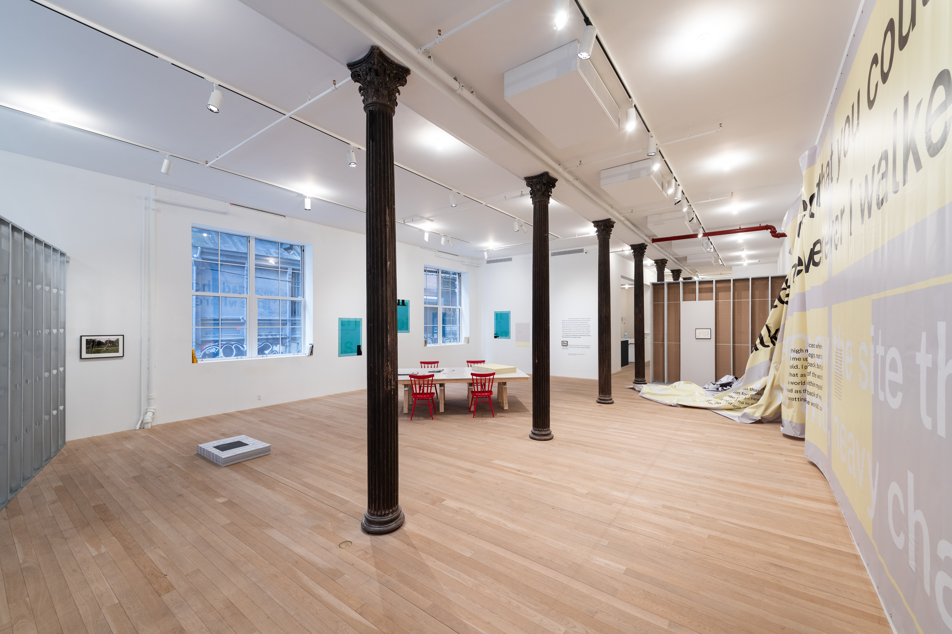 A color photograph of a gallery space featuring two large windows on the left and a row of dark metal columns in the center. A pile of grey posters, a table with red chairs, hanging framed images, and blue posters are visible in the space. A large grey and yellow vinyl partially hangs from the wall on the right, cascading down to the ground in the far right corner.