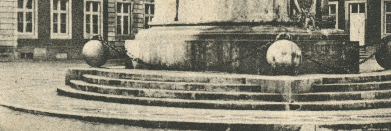 A black and white photograph of the base of a monument in a city square. Steps lead up to a pedastal surrounded by three large spheres connected by a metal chain.