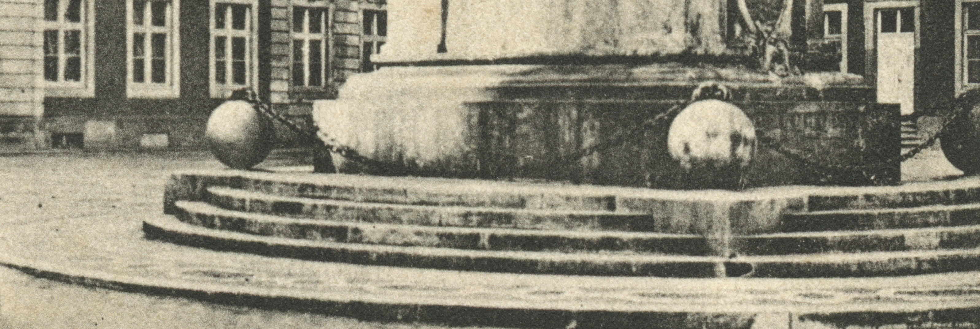 A black and white photograph of the base of a monument in a city square. Steps lead up to a pedastal surrounded by three large spheres connected by a metal chain.