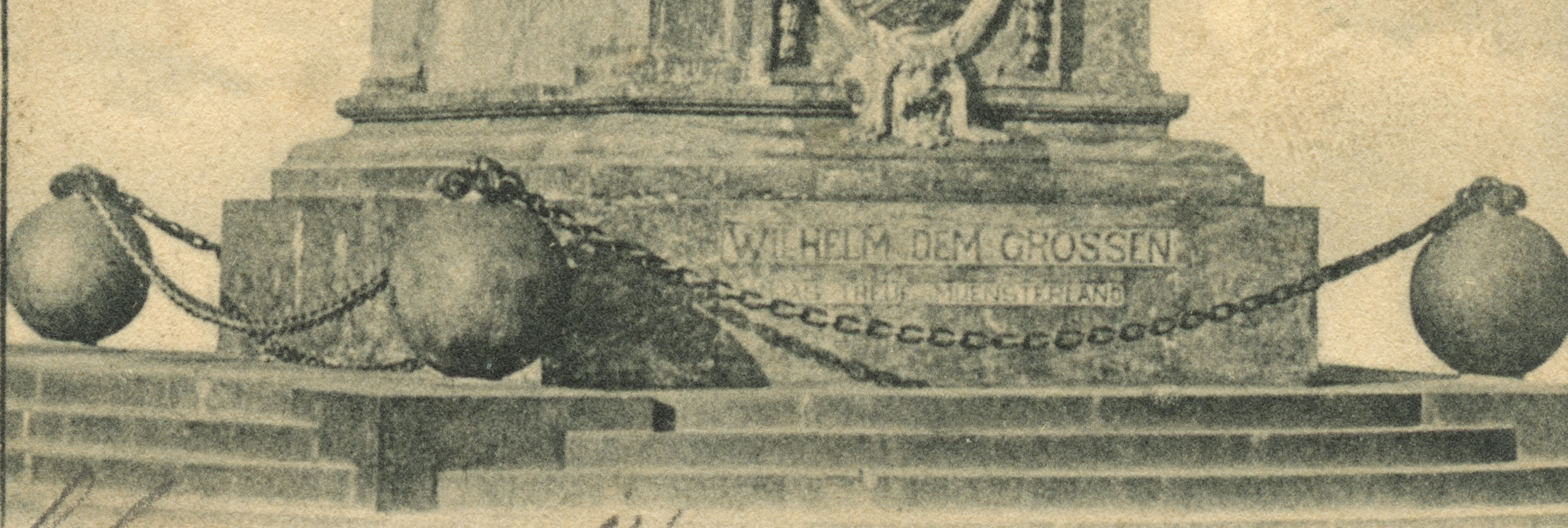 A black and white photograph of the base of a monument in a city square. Steps lead up to a pedastal surrounded by three large spheres connected by a metal chain.