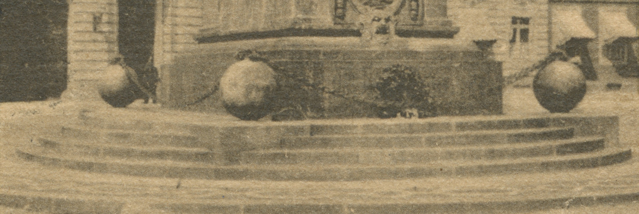 A black and white photograph of the base of a monument in a city square. Steps lead up to a pedastal surrounded by three large spheres connected by a metal chain.