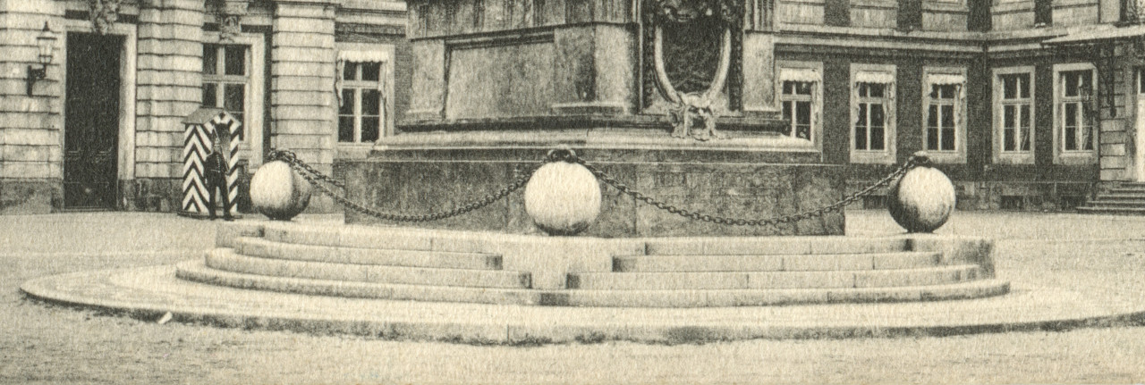 A black and white photograph of the base of a monument in a city square. Steps lead up to a pedastal surrounded by three large spheres connected by a metal chain.
