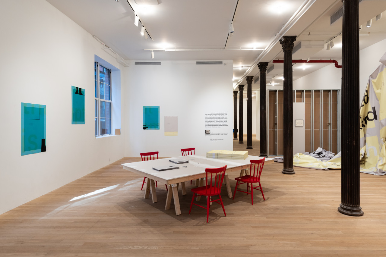 A color photograph of a gallery space. Blue posters hang from the left and rear walls. A large grey table with red chairs is in the center with stacks of yellow and grey posters. To the right of a row of dark metal columns, a large piece of grey and yellow vinyl partially hangs from the wall.