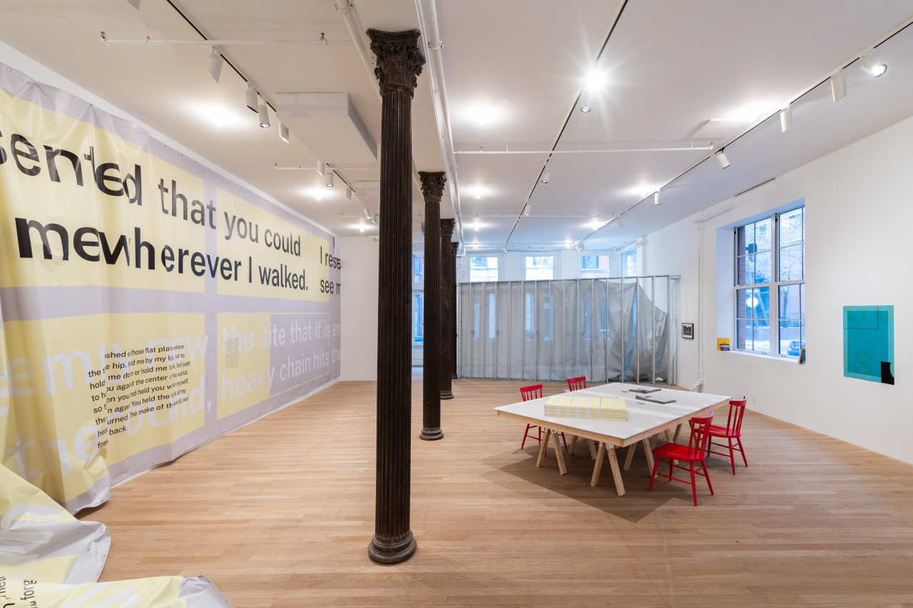 A color photograph of a gallery space. On the left, a large piece of grey and white vinyl hangs from the wall. A row of dark metal columns occupies the center of the space, extending back to a perpendicular section of grey metal scaffolding. On the right of the space there is a large grey table with four red chairs. The table holds a stack of yellow posters and two smaller stacks of grey handouts. The wall on the right has a large window and a blue poster.