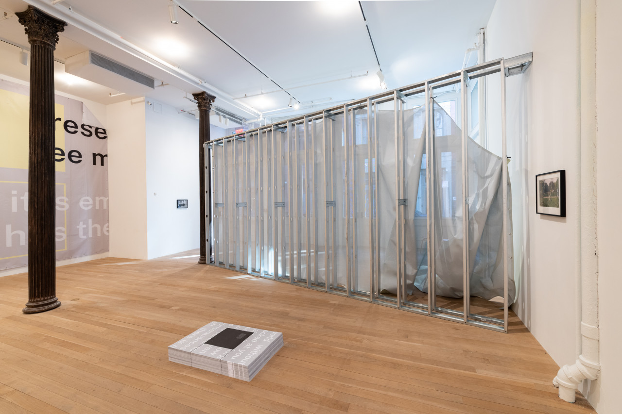 A color photograph of a gallery space. A large piece of grey and yellow vinyl hangs from the wall on the left, perpendicular to grey metal scaffolding with semi-transparent fabric on one side. A stack of grey a white posters are placed on the ground directly in front of the scaffold.