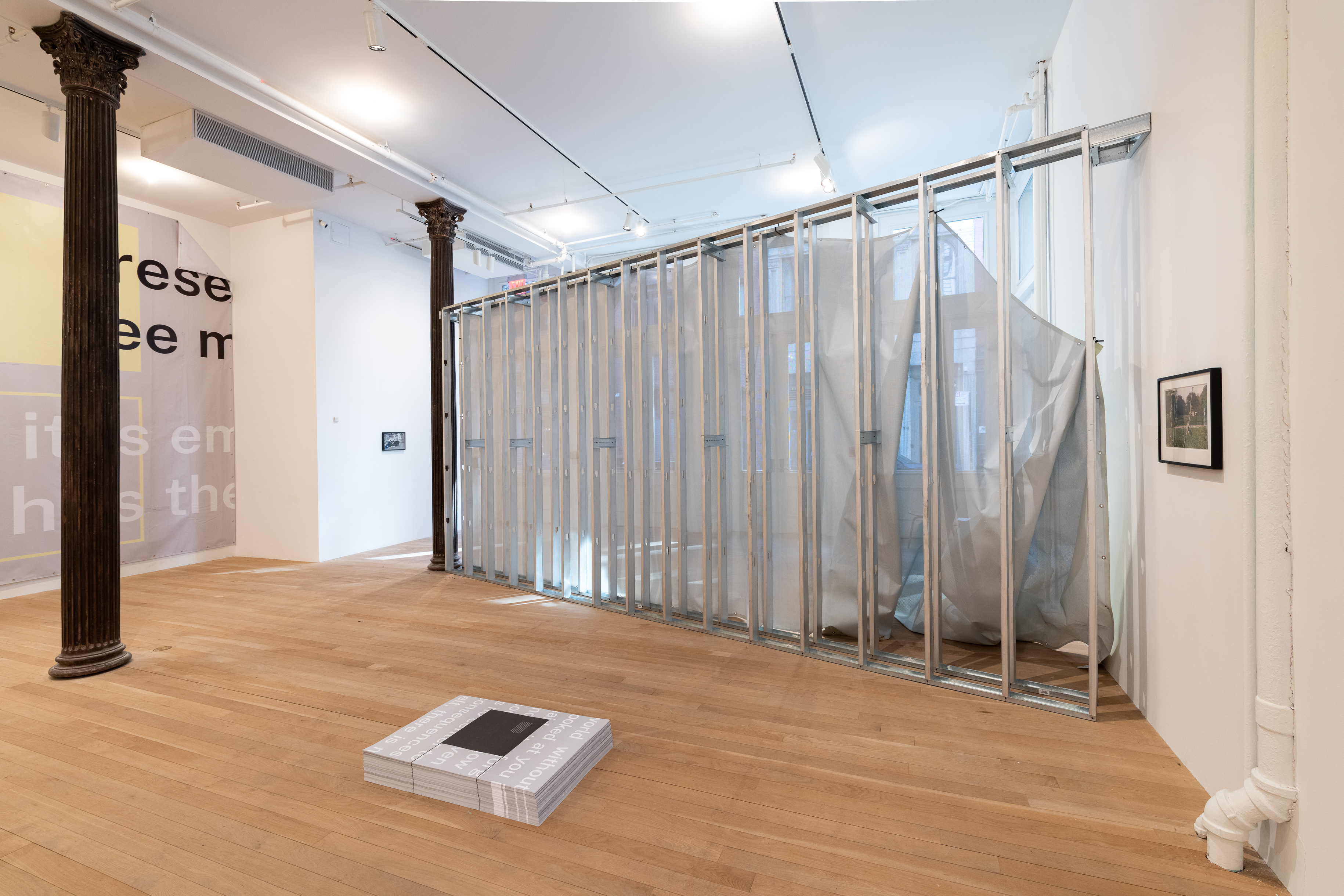 A color photograph of a gallery space. A large piece of grey and yellow vinyl hangs from the wall on the left, perpendicular to grey metal scaffolding with semi-transparent fabric on one side. A stack of grey a white posters are placed on the ground directly in front of the scaffold.