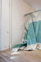 A color photograph of a grey and blue piece of semi-transparent fabric, partially hung from a grey metal scaffold in a gallery space.