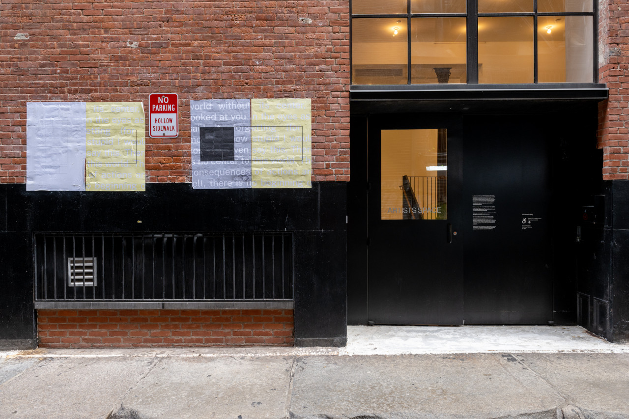 A color photograph of an alleyway. Two sets of grey and yellow posters hang on the brick surface, next to a large black doorway.