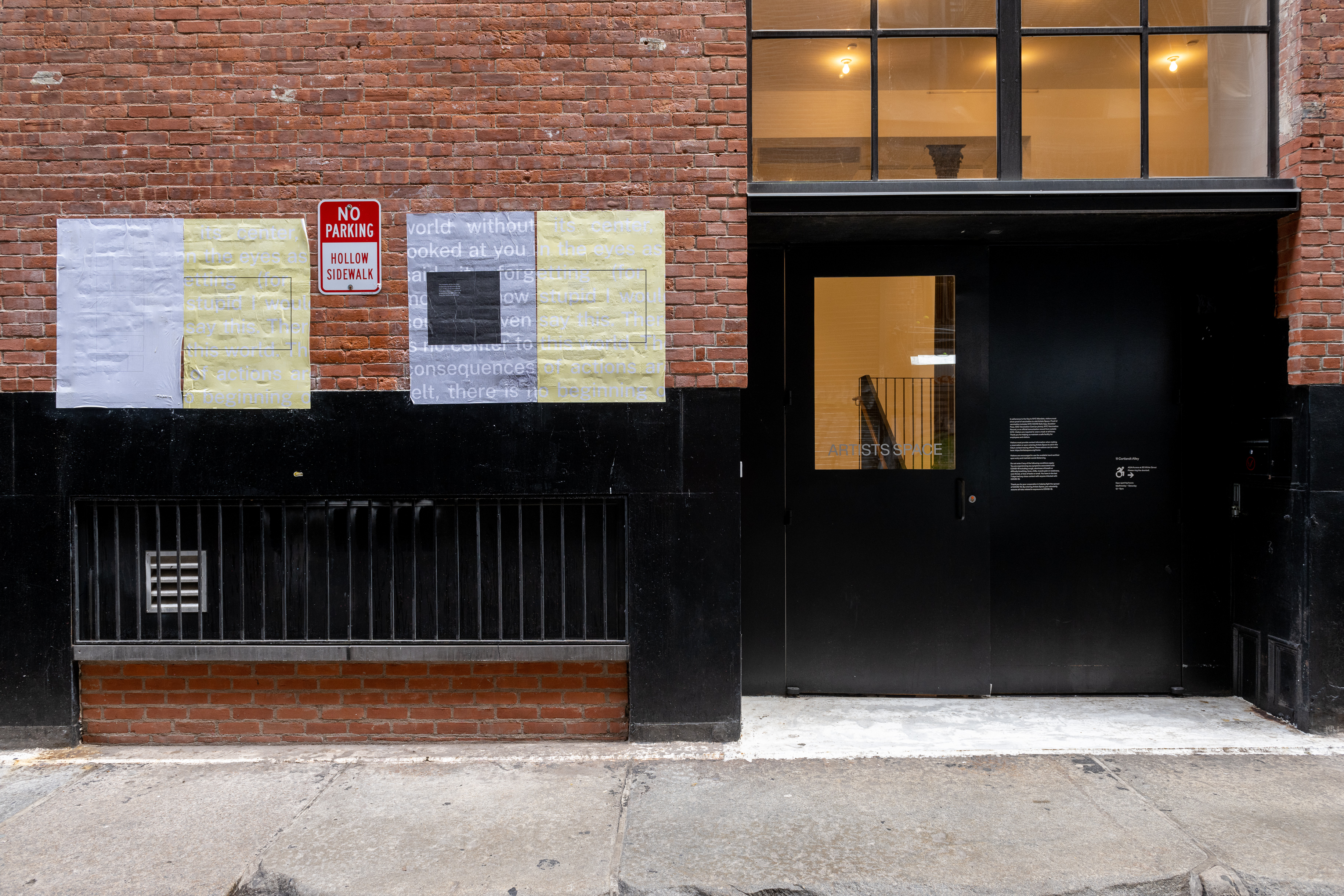 A color photograph of an alleyway. Two sets of grey and yellow posters hang on the brick surface, next to a large black doorway.