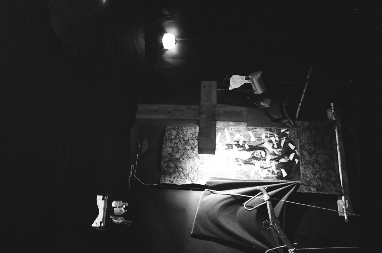 A painting hanging next to a bag behind a cross with light shining on it. A lighted ball is seen in one corner and lighted bottles in a box are seen in another corner.