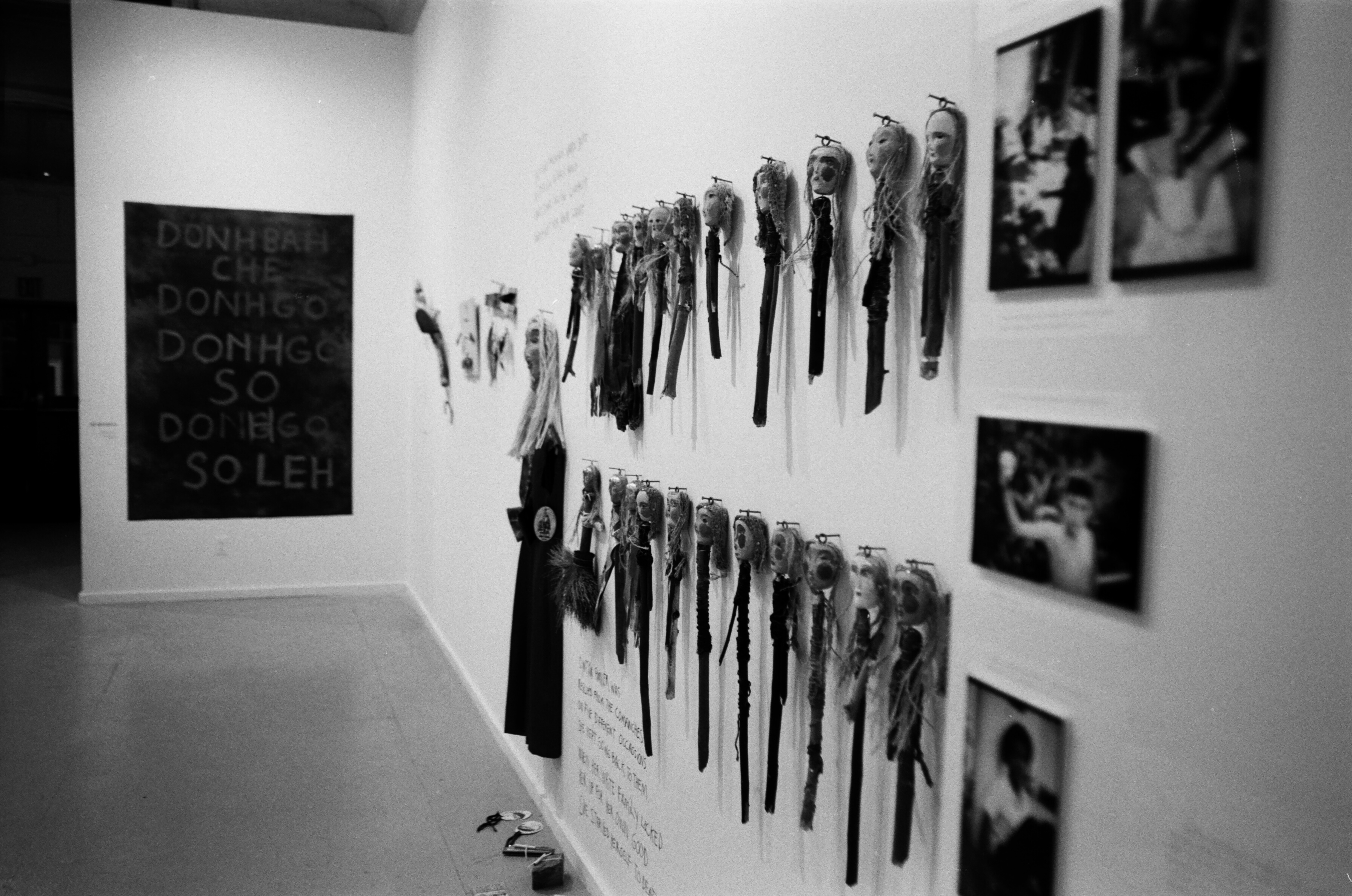 Dolls with photos adjacaent to them hangon the wall on protruding nails and black text is also visible around them. An object can be seen on the floor and a text-based painting is on a wall adjacent to the dolls in this white-walled gallery setting.