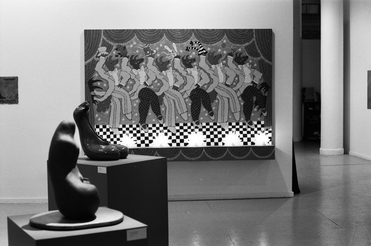 A painting appears on the wall of figures dancing and two sculptures can be seen on pedestals in the foreground in a gallery setting.