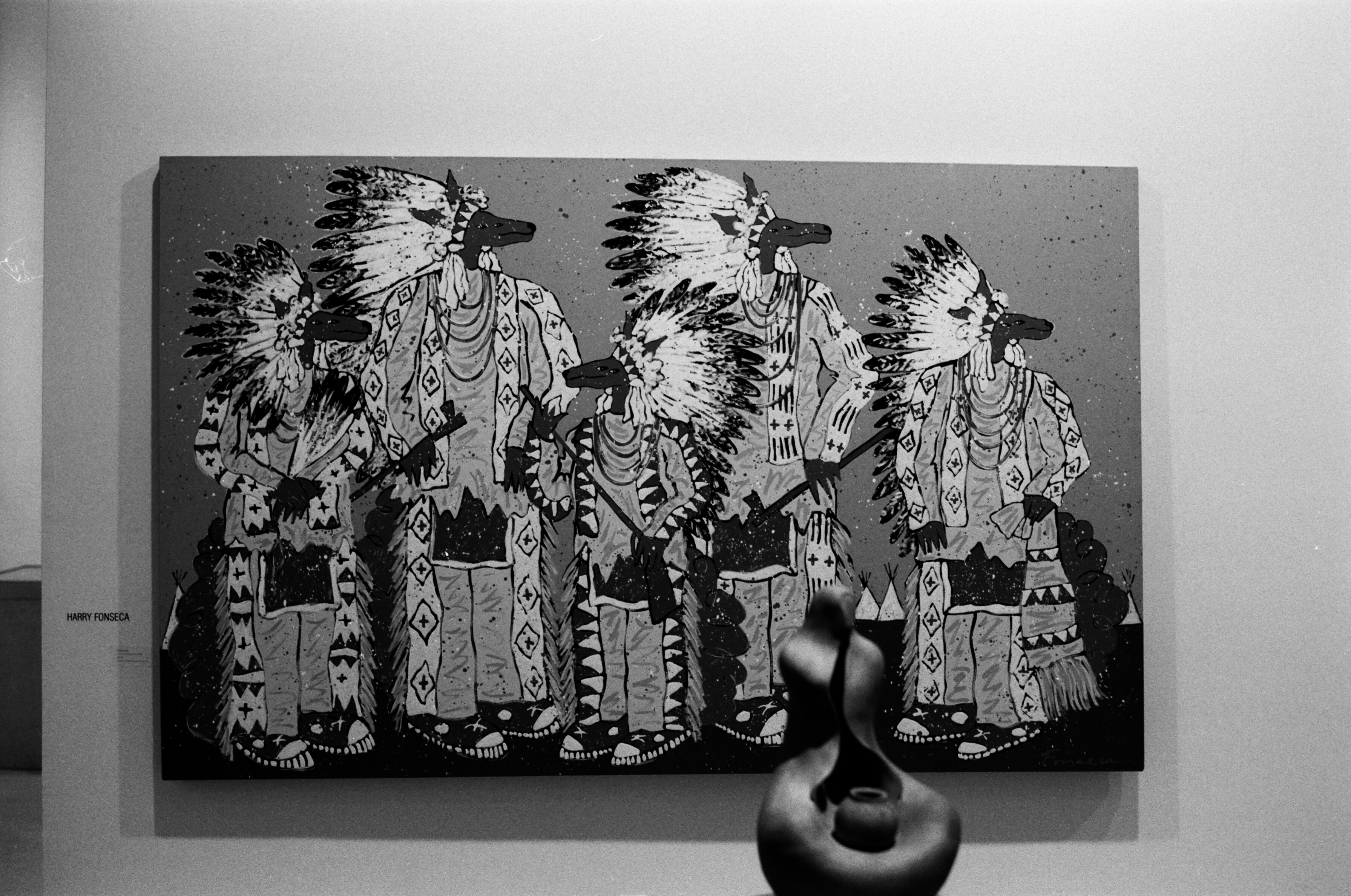 Painting of figures with Native American headdresses on a white gallery wall with a sculpture appearing in front of it.