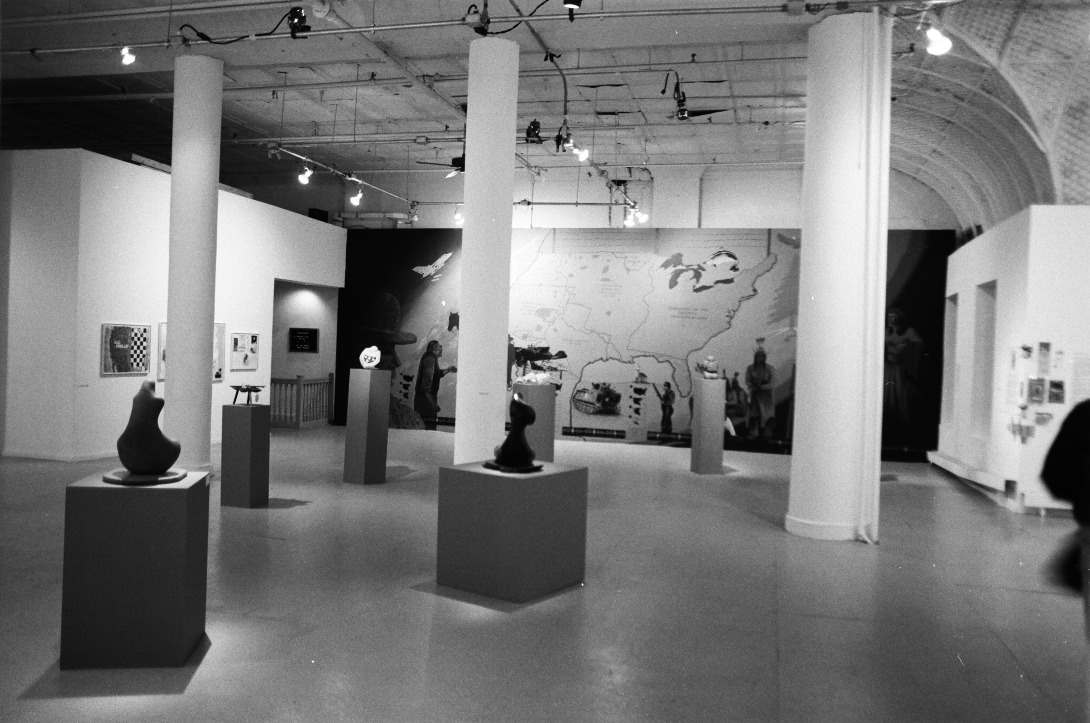 A number of sculptures appear on pedestals with paintings on walls behind them with columns bisecting the image in a gallery setting.