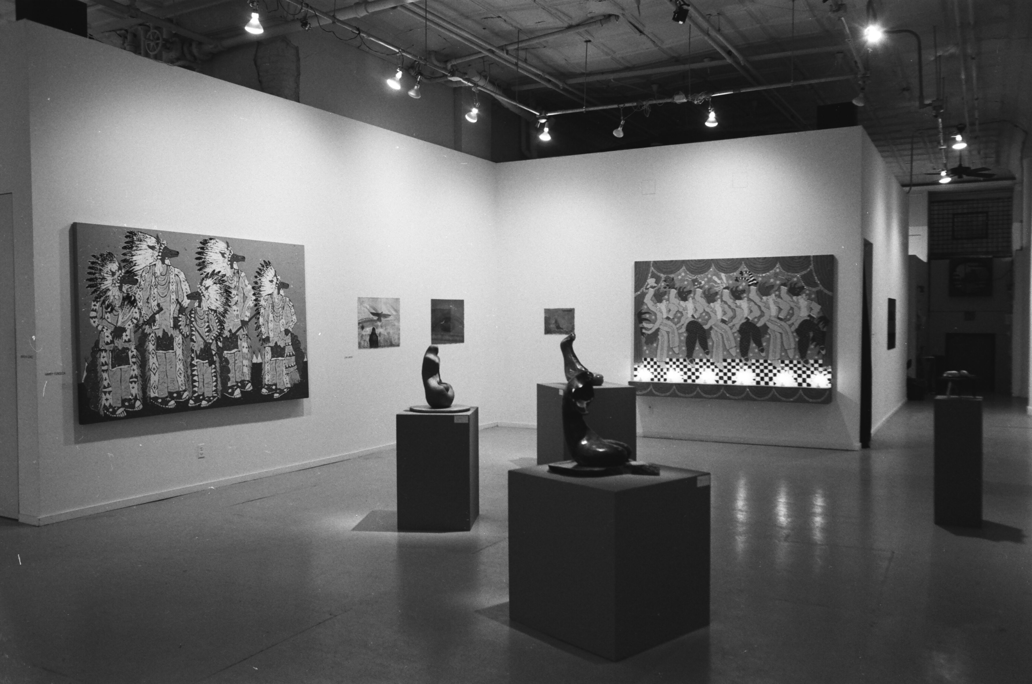 Dark sculptural elements appear lighted on boxes in the center of the image with paintings on the gallery walls behind them. A corridor is seen to the right.