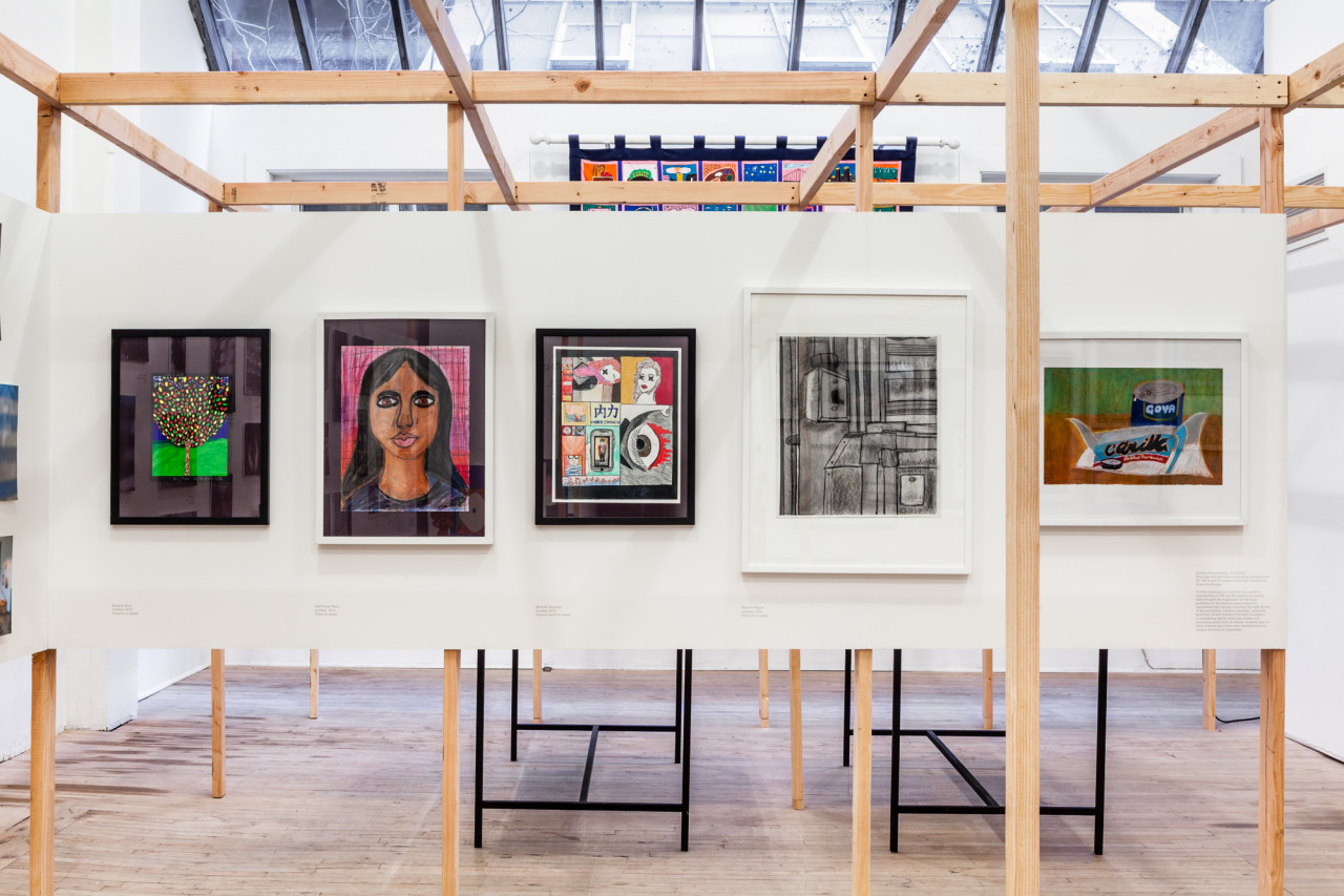 Five framed drawings hang on white panel, suspended on a wooden frame that extends into and across the room.