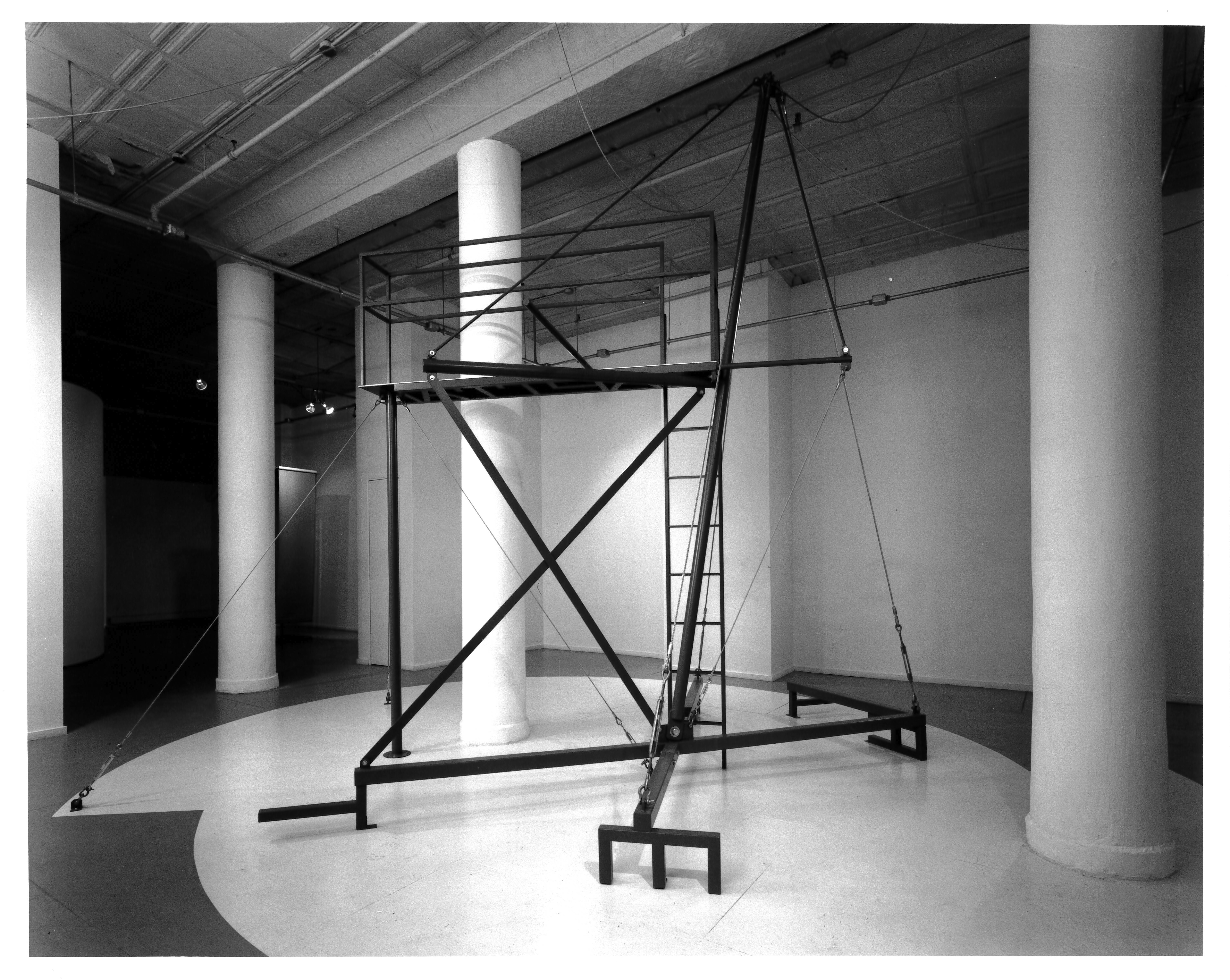 A black and white photograph of a tall metal sculpture in an otherwise empty room, surrounded by white columns. The sculpture has a tall platform with a narrow ladder and has several support wires.