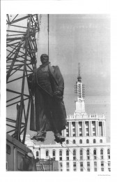 A metal statue is being lifted from its platform by a crane via ropes around its neck and feet. A large building is visible behind it.